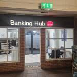 The new Mumbles Banking Hub entrance, with an illuminated black and white "Banking Hub" sign featuring the red Post Office logo above a glass door, set into a brick interior wall with bilingual Welsh and English signage and dog food displayed on shelving to the right