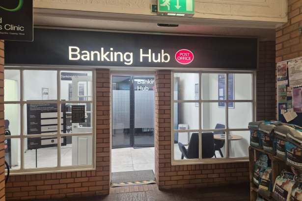 The new Mumbles Banking Hub entrance, with an illuminated black and white "Banking Hub" sign featuring the red Post Office logo above a glass door, set into a brick interior wall with bilingual Welsh and English signage and dog food displayed on shelving to the right