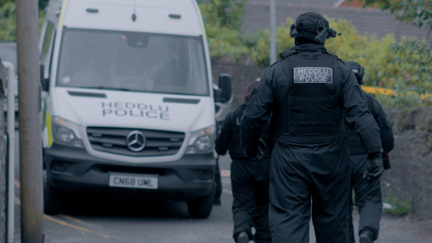 Two South Wales Police officers in black tactical clothing and protective vests with "Heddlu Police" badges walk towards a marked Mercedes Sprinter police van during a dawn raid in Swansea