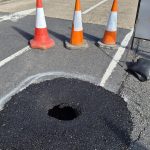 A large hole in the surface of Lando Road in Pembrey, surrounded by traffic cones and spray-painted white markings, showing the extent of the sinkhole beneath the road surface