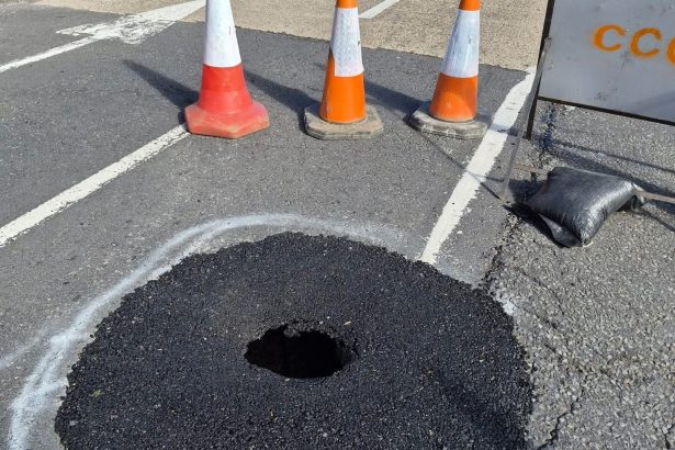A large hole in the surface of Lando Road in Pembrey, surrounded by traffic cones and spray-painted white markings, showing the extent of the sinkhole beneath the road surface