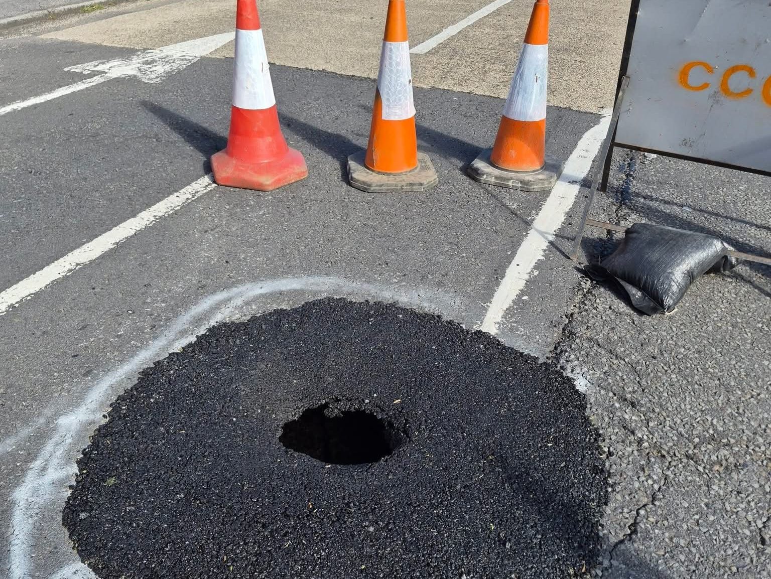 A large hole in the surface of Lando Road in Pembrey, surrounded by traffic cones and spray-painted white markings, showing the extent of the sinkhole beneath the road surface