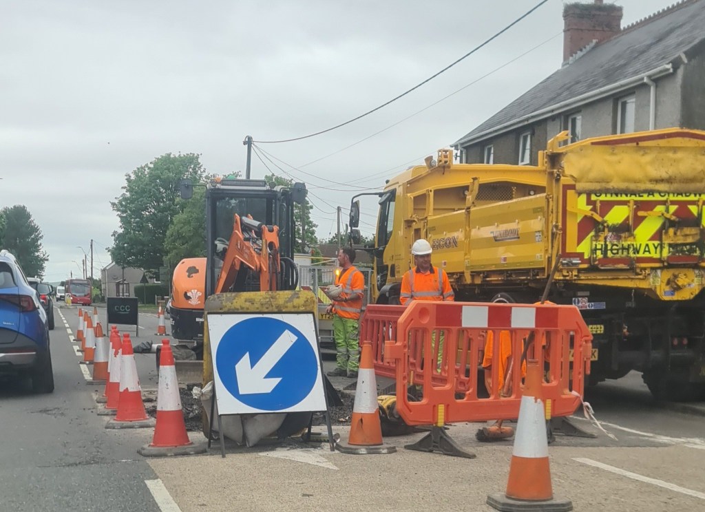 Carmarthenshire County Council highway maintenance workers and vehicles on Lando Road in Pembrey, with orange barriers, cones and a yellow excavator visible during emergency repair works