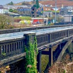 Cymmer Bridge, a stone arch bridge with concrete balustrade railings and ivy growing on the central pier, spanning a wooded valley with the village of Cymmer behind it, including the fire station with its red doors visible alongside parked ambulances.