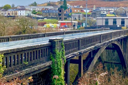 Cymmer Bridge, a stone arch bridge with concrete balustrade railings and ivy growing on the central pier, spanning a wooded valley with the village of Cymmer behind it, including the fire station with its red doors visible alongside parked ambulances.