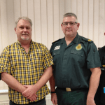 Five men stand together in a row indoors at a Save a Life Cymru reunion event, with two wearing the dark green uniform of the Welsh Ambulance Service, one in a yellow checked shirt holding a Bystander Commendation Certificate, one in a black Mitie security fleece, and one in a blue shirt holding another certificate