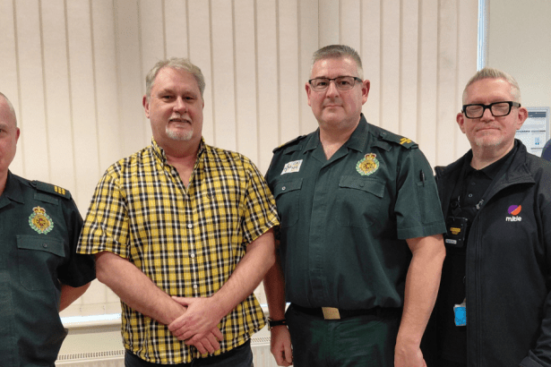Five men stand together in a row indoors at a Save a Life Cymru reunion event, with two wearing the dark green uniform of the Welsh Ambulance Service, one in a yellow checked shirt holding a Bystander Commendation Certificate, one in a black Mitie security fleece, and one in a blue shirt holding another certificate