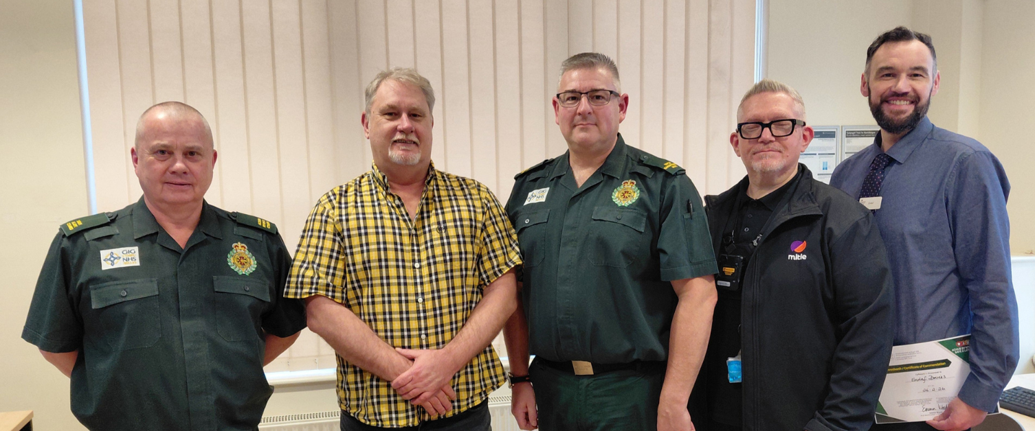 Five men stand together in a row indoors at a Save a Life Cymru reunion event, with two wearing the dark green uniform of the Welsh Ambulance Service, one in a yellow checked shirt holding a Bystander Commendation Certificate, one in a black Mitie security fleece, and one in a blue shirt holding another certificate