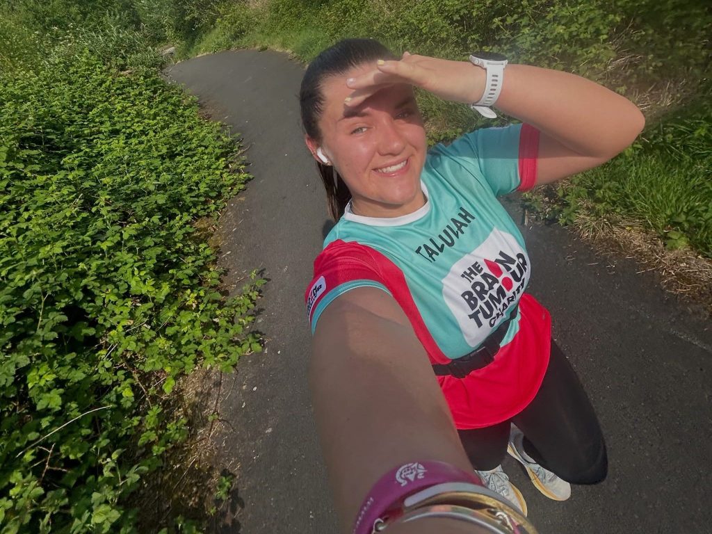 Tallulah Chalstrey takes a selfie on a training run, wearing a teal and red Brain Tumour Charity vest with her name on it, shielding her eyes from the sun on a greenery-lined path.