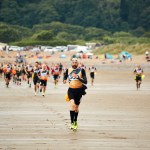 A large group of competitors in wetsuits and race vests run across a wide sandy beach during the Gritty Rascals Swimrun Gower event, with trees and parked vehicles visible in the background.