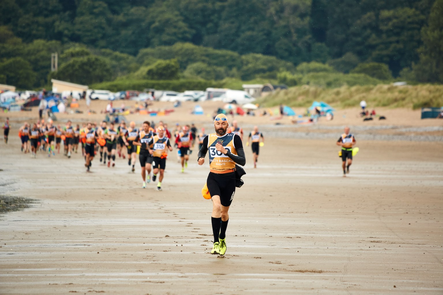 A large group of competitors in wetsuits and race vests run across a wide sandy beach during the Gritty Rascals Swimrun Gower event, with trees and parked vehicles visible in the background.