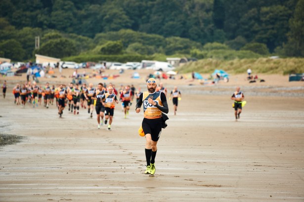 A large group of competitors in wetsuits and race vests run across a wide sandy beach during the Gritty Rascals Swimrun Gower event, with trees and parked vehicles visible in the background.