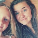 A selfie of two young girls smiling at the camera — Lexi May Hopkin on the left with blonde hair and blue eyes, and a younger Tallulah Chalstrey on the right with longer brown hair.