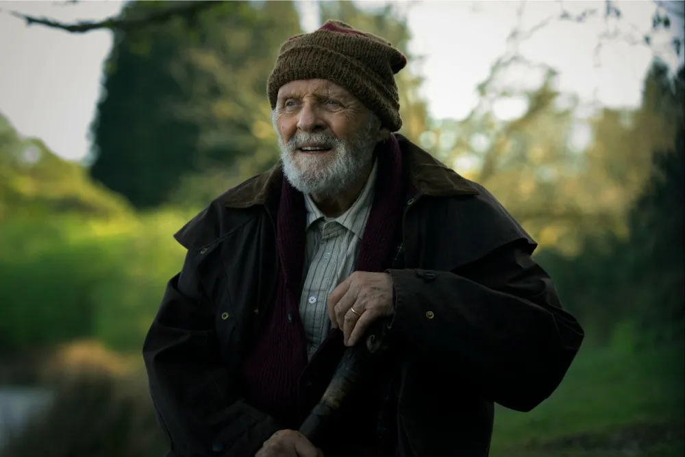 Sir Anthony Hopkins as the grandfather character in A Visit to Grandpa's, wearing a woolly hat and dark coat and holding a walking stick in an outdoor rural setting with trees in the background