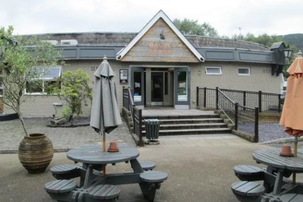 The exterior of the Bagle Brook pub restaurant in Baglan, showing the Bagle Brook sign above the entrance, outdoor picnic tables and parasols on a paved forecourt, with trees and hills visible in the background
