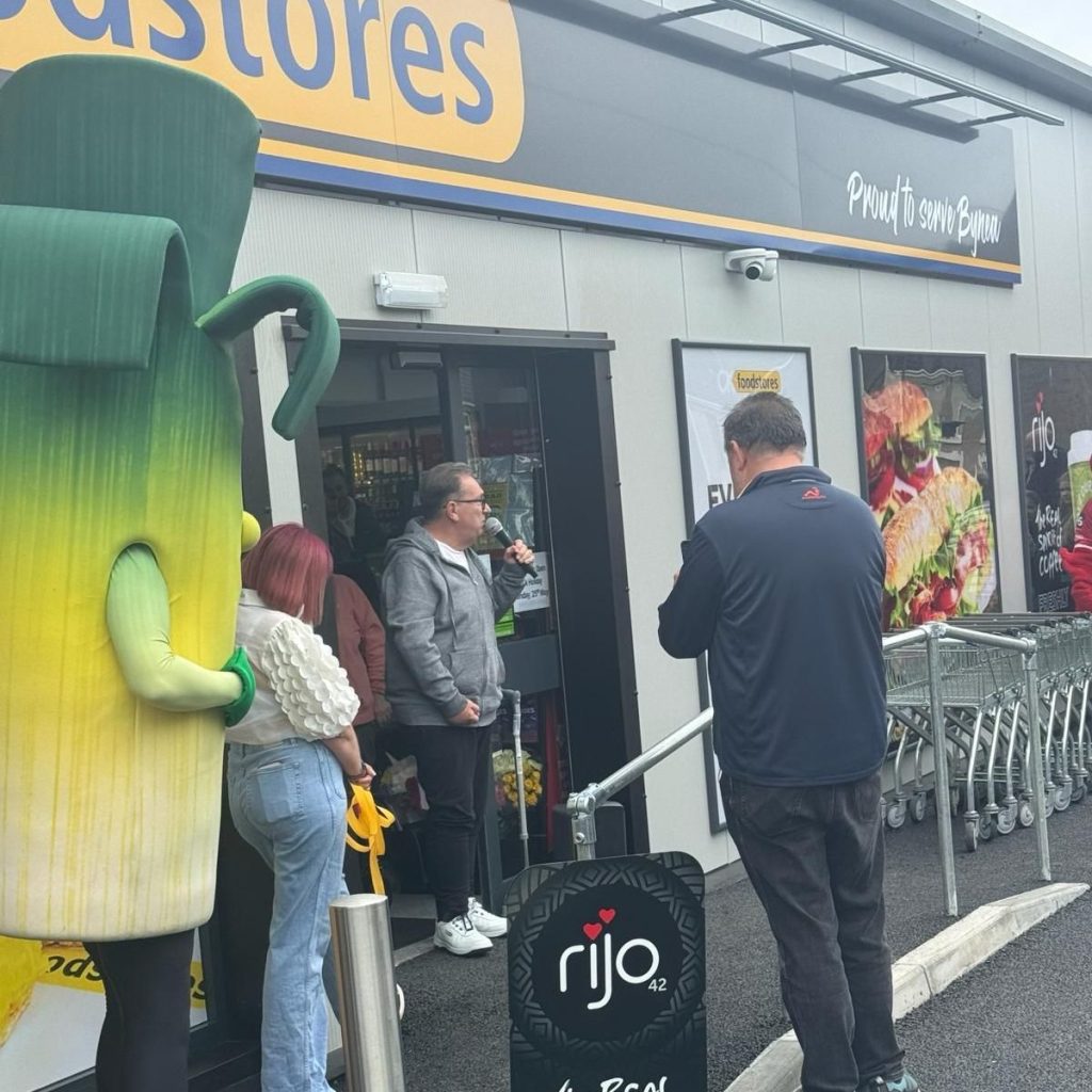 Kev Johns MBE speaks at the microphone outside the new CK Foodstores in Bynea, with the store's "Proud to serve Bynea" fascia visible above the entrance and a giant leek mascot to the left.