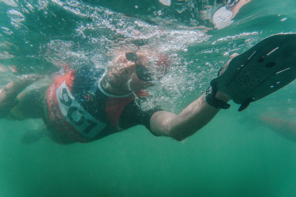 An underwater photograph of a competitor in a red and black Gritty Rascals race vest and goggles, captured mid-stroke in the green waters off the Gower coast.