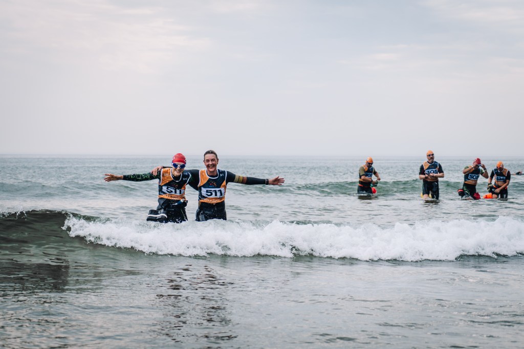Two male competitors in orange and black Gritty Rascals race vests stand arm-in-arm on a pebble beach, smiling at the camera, with the Mumbles lighthouse and rocky headland visible behind them.