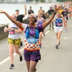Smiling female runner with her arms raised in celebration during a previous Mizuno Swansea Half Marathon, running along the Swansea Bay coastal path with fellow runners and spectators behind her.