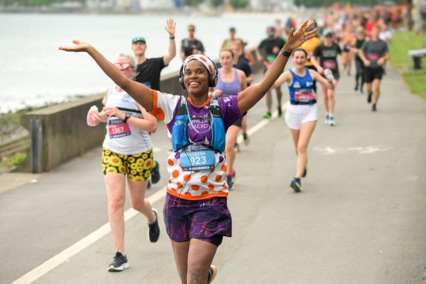 Smiling female runner with her arms raised in celebration during a previous Mizuno Swansea Half Marathon, running along the Swansea Bay coastal path with fellow runners and spectators behind her.