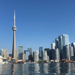The Toronto city skyline viewed from the water on a clear blue day, dominated by the CN Tower with the Rogers Centre stadium visible to the left and modern skyscrapers stretching across the waterfront
