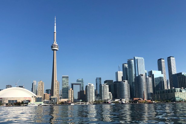 The Toronto city skyline viewed from the water on a clear blue day, dominated by the CN Tower with the Rogers Centre stadium visible to the left and modern skyscrapers stretching across the waterfront