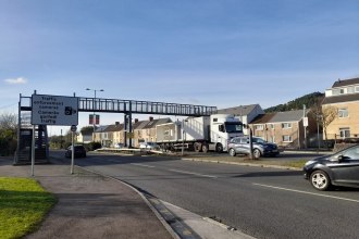 A view of Fabian Way in Swansea showing a dual carriageway with a pedestrian footbridge overhead, heavy goods vehicles and cars on the road, and a bilingual traffic enforcement camera warning sign in Welsh and English