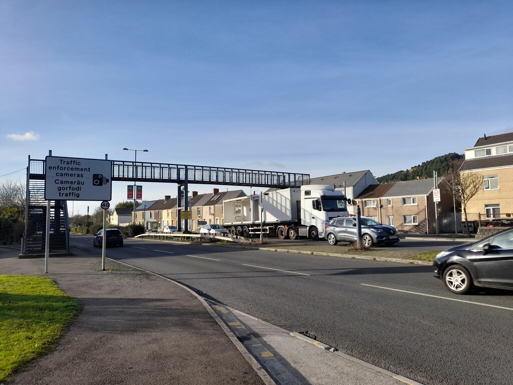 A view of Fabian Way in Swansea showing a dual carriageway with a pedestrian footbridge overhead, heavy goods vehicles and cars on the road, and a bilingual traffic enforcement camera warning sign in Welsh and English