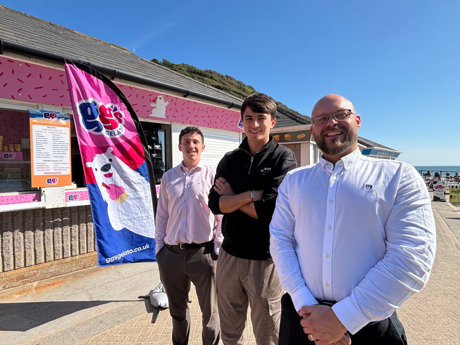 Tom Mogridge, Toby Link-Jones and James Smale-Davies stand together on a sunny seafront promenade outside the pink-painted GG's Gelato kiosk, with a branded GG's Gelato flag and menu board visible behind them