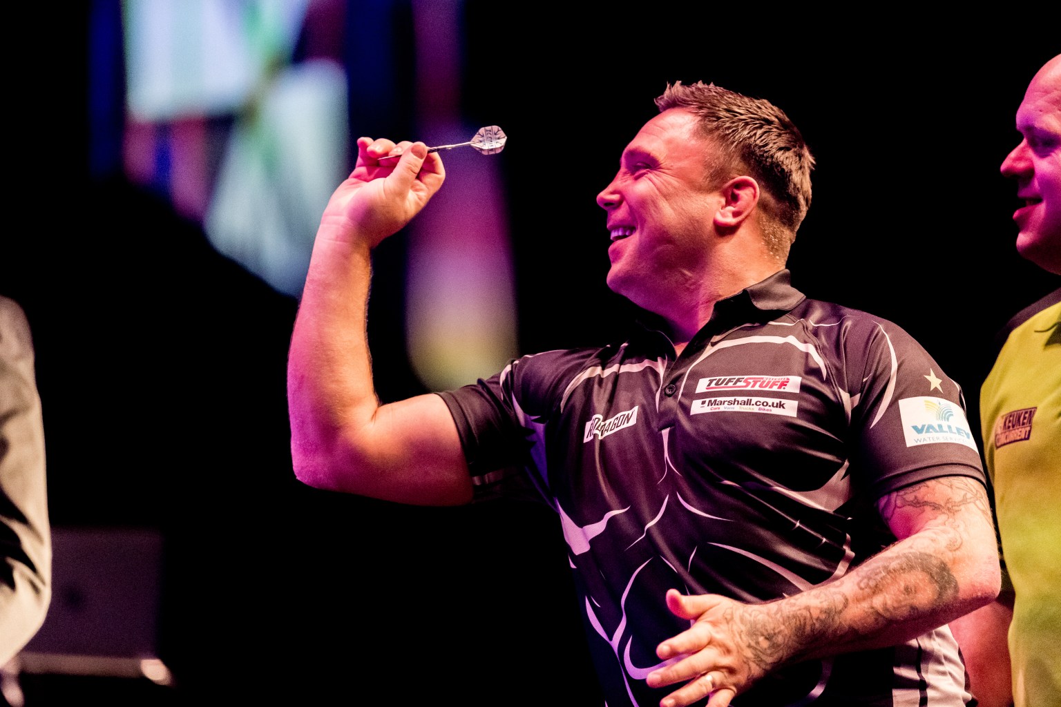 Gerwyn Price, wearing a black darts shirt, raises his arm to throw a dart while smiling during a darts event