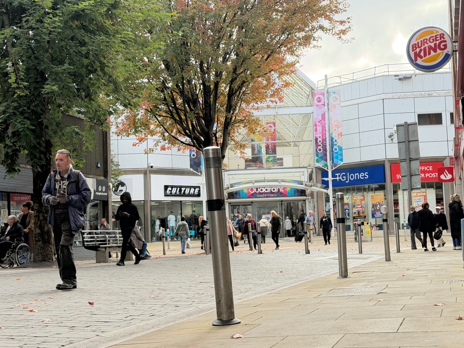 A view of the TGJones store at the entrance to the Quadrant Shopping Centre in Swansea, with the chain's blue signage visible to the right of the centre's main entrance, alongside a Post Office sign, Burger King logo and shoppers walking in the autumn pedestrianised area