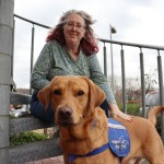 Emma Reardon, a woman with red-streaked grey hair and glasses, sits on steps outdoors with her hand resting on her Red Fox Labrador Jeff, who is wearing a blue Support Dog vest and looking directly at the camera.