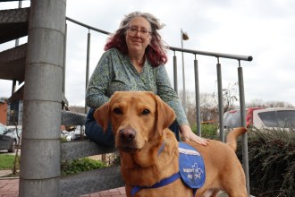 Emma Reardon, a woman with red-streaked grey hair and glasses, sits on steps outdoors with her hand resting on her Red Fox Labrador Jeff, who is wearing a blue Support Dog vest and looking directly at the camera.