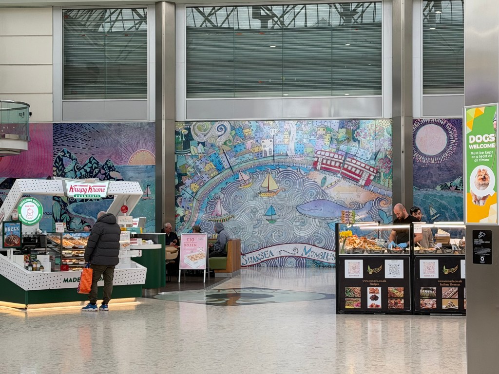 Interior view of the Quadrant Shopping Centre showing a Krispy Kreme kiosk on the left, an O'Snack food display on the right, and a large mural depicting Swansea Bay and Mumbles in the background, occupying what was the entrance to the Debenhams store.