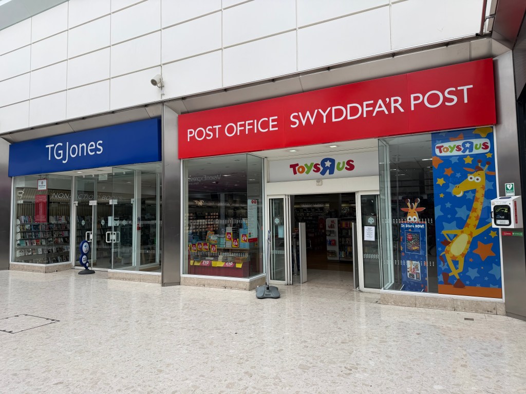 A close-up of the TGJones storefront inside the Quadrant Shopping Centre in Swansea, showing the chain's blue signage to the left, a red Post Office sign in Welsh and English ("Swyddfa'r Post") in the centre and a Toys R Us giraffe display to the right