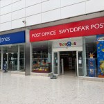 A close-up of the TGJones storefront inside the Quadrant Shopping Centre in Swansea, showing the chain's blue signage to the left, a red Post Office sign in Welsh and English ("Swyddfa'r Post") in the centre and a Toys R Us giraffe display to the right