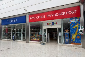 A close-up of the TGJones storefront inside the Quadrant Shopping Centre in Swansea, showing the chain's blue signage to the left, a red Post Office sign in Welsh and English ("Swyddfa'r Post") in the centre and a Toys R Us giraffe display to the right