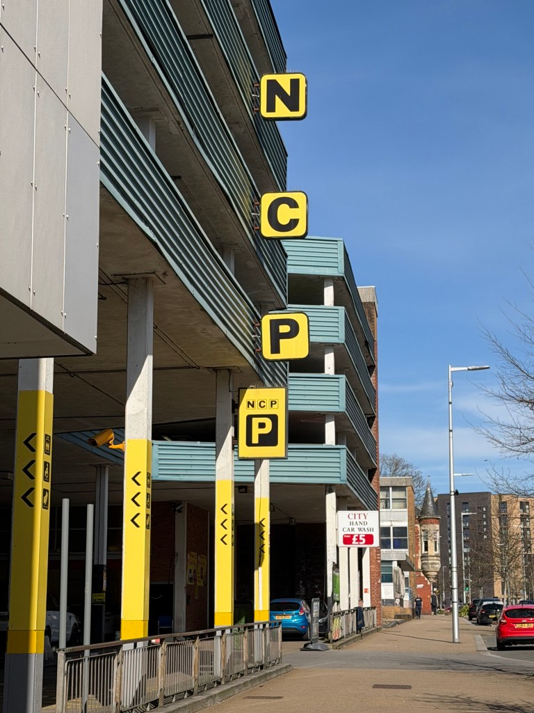 A street-level view of the NCP Orchard Street Car Park in Swansea showing the large yellow NCP lettering against the blue-fronted multi-storey structure, with parked cars and a City Hand Car Wash sign visible nearby
