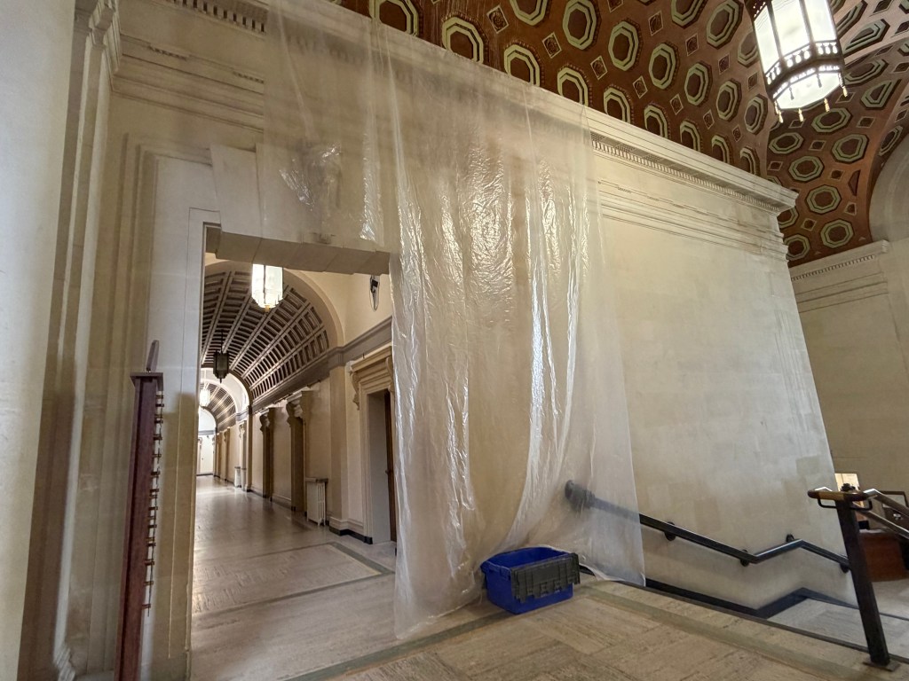 A long sheet of clear plastic sheeting hangs from the ornate octagonal coffered ceiling of Swansea Guildhall down to a blue plastic bucket positioned on the steps, with a vaulted corridor visible through an archway to the left