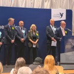 Six newly elected Members of the Senedd stand on stage at the Sir Gaerfyrddin count, with Plaid Cymru's Cefin Campbell standing at a wooden podium delivering remarks and Reform UK's Gareth Beer standing behind him. Lined up to one side in order of election are Plaid Cymru's Nerys Evans, Reform UK's Carmelo Colasanto, Plaid Cymru's Adam Price and Reform UK's Sarah Edwards.