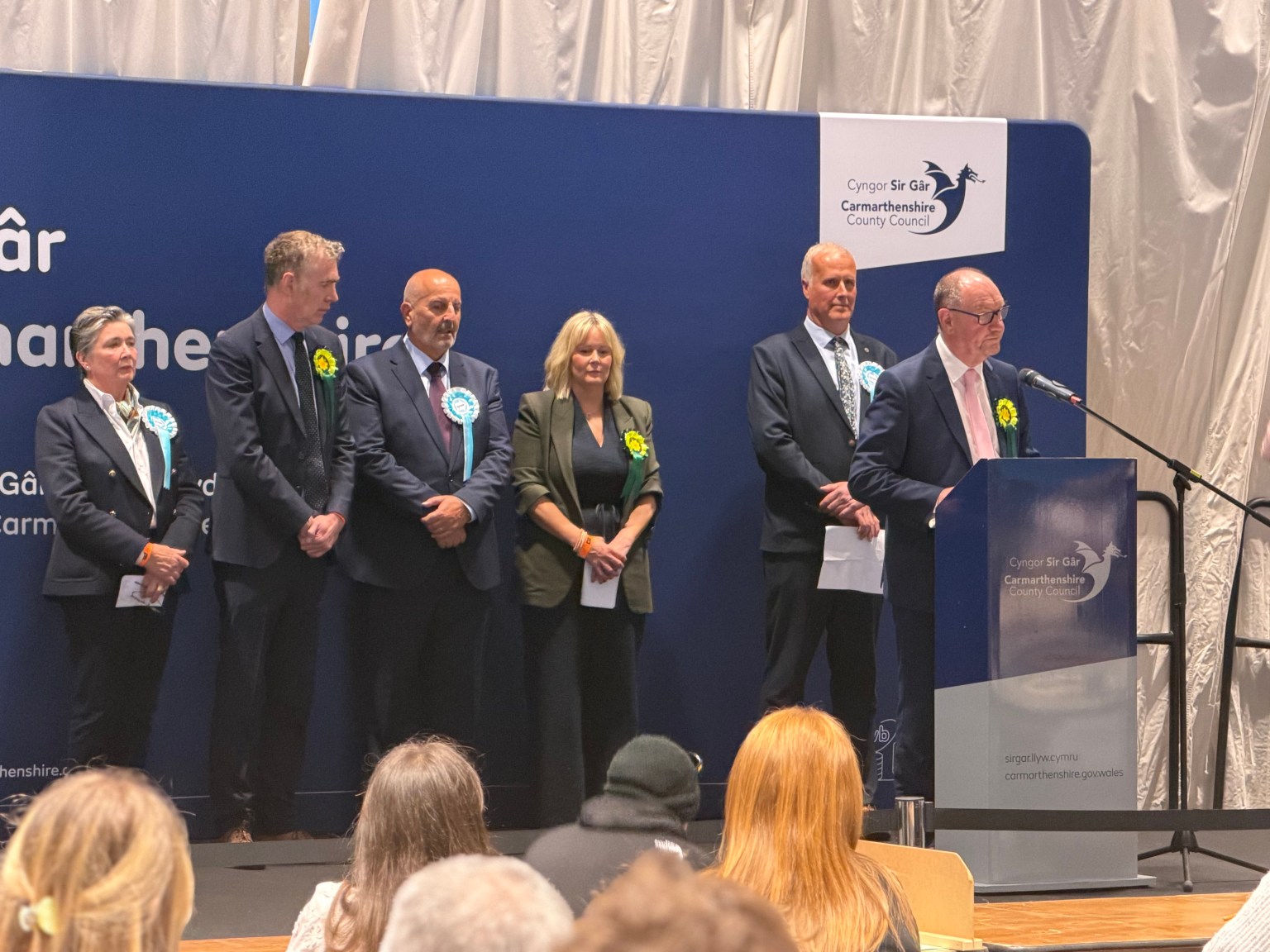 Six newly elected Members of the Senedd stand on stage at the Sir Gaerfyrddin count, with Plaid Cymru's Cefin Campbell standing at a wooden podium delivering remarks and Reform UK's Gareth Beer standing behind him. Lined up to one side in order of election are Plaid Cymru's Nerys Evans, Reform UK's Carmelo Colasanto, Plaid Cymru's Adam Price and Reform UK's Sarah Edwards.
