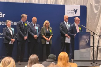Six newly elected Members of the Senedd stand on stage at the Sir Gaerfyrddin count, with Plaid Cymru's Cefin Campbell standing at a wooden podium delivering remarks and Reform UK's Gareth Beer standing behind him. Lined up to one side in order of election are Plaid Cymru's Nerys Evans, Reform UK's Carmelo Colasanto, Plaid Cymru's Adam Price and Reform UK's Sarah Edwards.