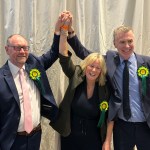 Three Plaid Cymru politicians celebrate at the Sir Gaerfyrddin Senedd count, all wearing yellow and green Plaid Cymru rosettes — Cefin Campbell on the left in a navy suit and pink tie, Nerys Evans centre in a black dress, and Adam Price on the right in a navy suit and tie — with their hands clasped together and raised in the air