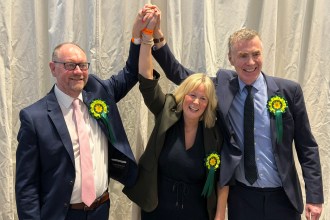 Three Plaid Cymru politicians celebrate at the Sir Gaerfyrddin Senedd count, all wearing yellow and green Plaid Cymru rosettes — Cefin Campbell on the left in a navy suit and pink tie, Nerys Evans centre in a black dress, and Adam Price on the right in a navy suit and tie — with their hands clasped together and raised in the air