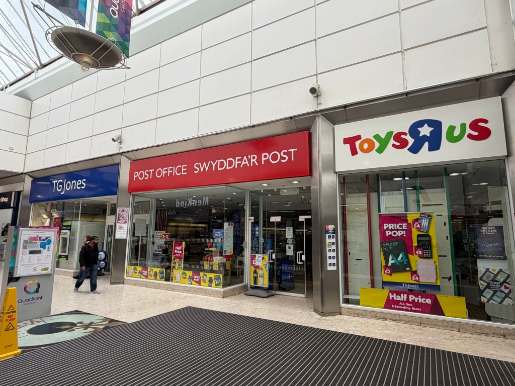 Three shopfronts inside the Quadrant Shopping Centre — a TGJones store on the left, the Post Office and Swyddfa'r Post in the middle, and a Toys R Us store on the right, with two shoppers walking past on a polished tiled floor.