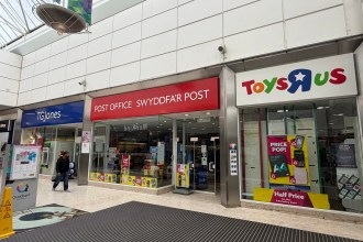 Three shopfronts inside the Quadrant Shopping Centre — a TGJones store on the left, the Post Office and Swyddfa'r Post in the middle, and a Toys R Us store on the right, with two shoppers walking past on a polished tiled floor.