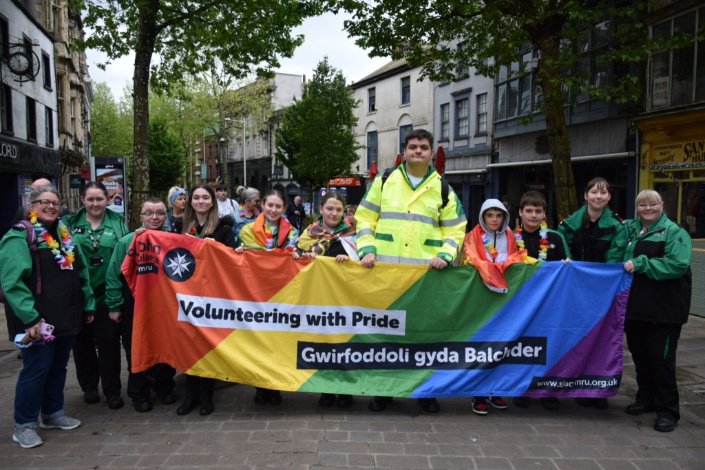 A group of St John Ambulance volunteers of varying ages march in the Swansea Pride 2026 parade carrying a bilingual Welsh and English banner reading Volunteering with Pride and Gwirfoddoli gyda Balchder.