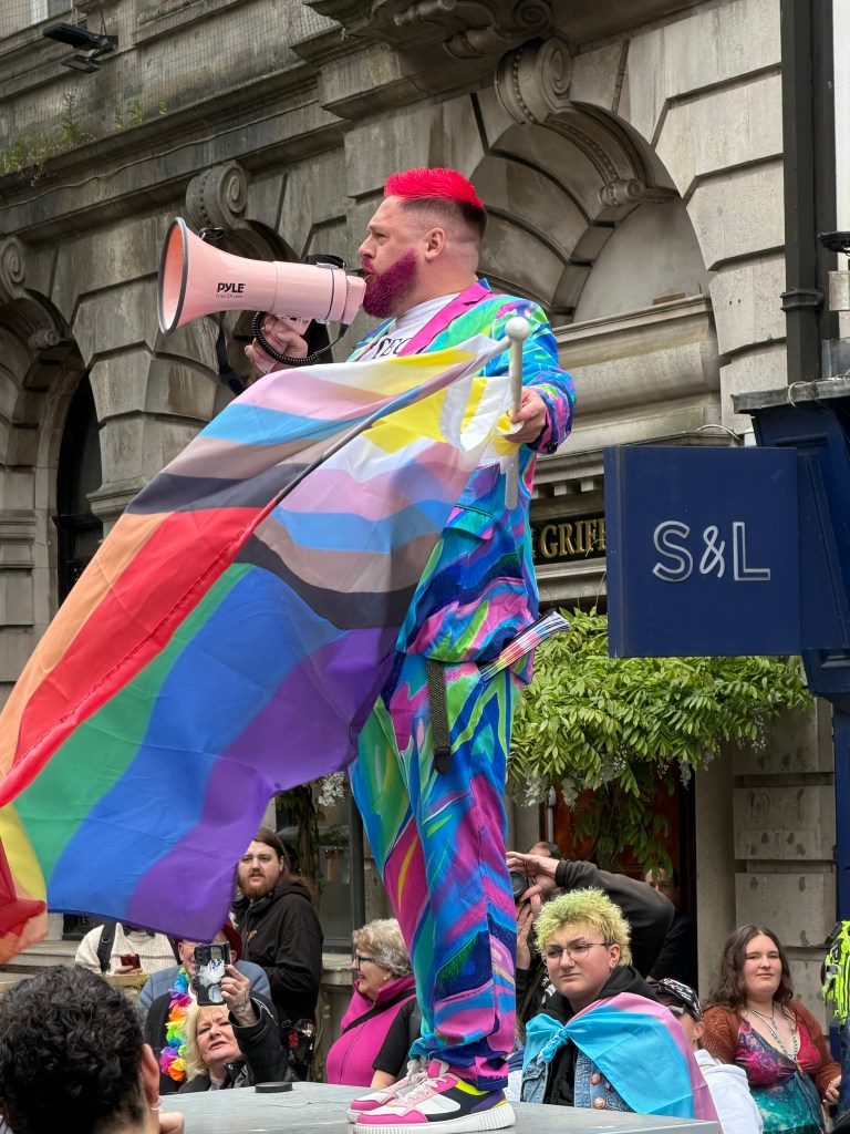 Pride host Jamie J, wearing a colourful patterned suit and red hair, stands elevated above a large crowd on Wind Street speaking into a megaphone with a Progress Pride flag, before the Swansea Pride 2026 parade begins.