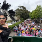 Drag queen Chae with a C, wearing a black sequinned gown and elaborate feathered headdress, stands on the upper deck of the Swansea Pride open-top bus looking back over the parade, with hundreds of marchers carrying flags and banners visible in the street behind her.