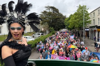 Drag queen Chae with a C, wearing a black sequinned gown and elaborate feathered headdress, stands on the upper deck of the Swansea Pride open-top bus looking back over the parade, with hundreds of marchers carrying flags and banners visible in the street behind her.